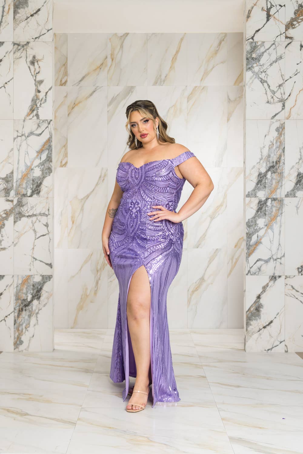 A woman wearing a purple off-shoulder dress with a slit in front against a marble wall.