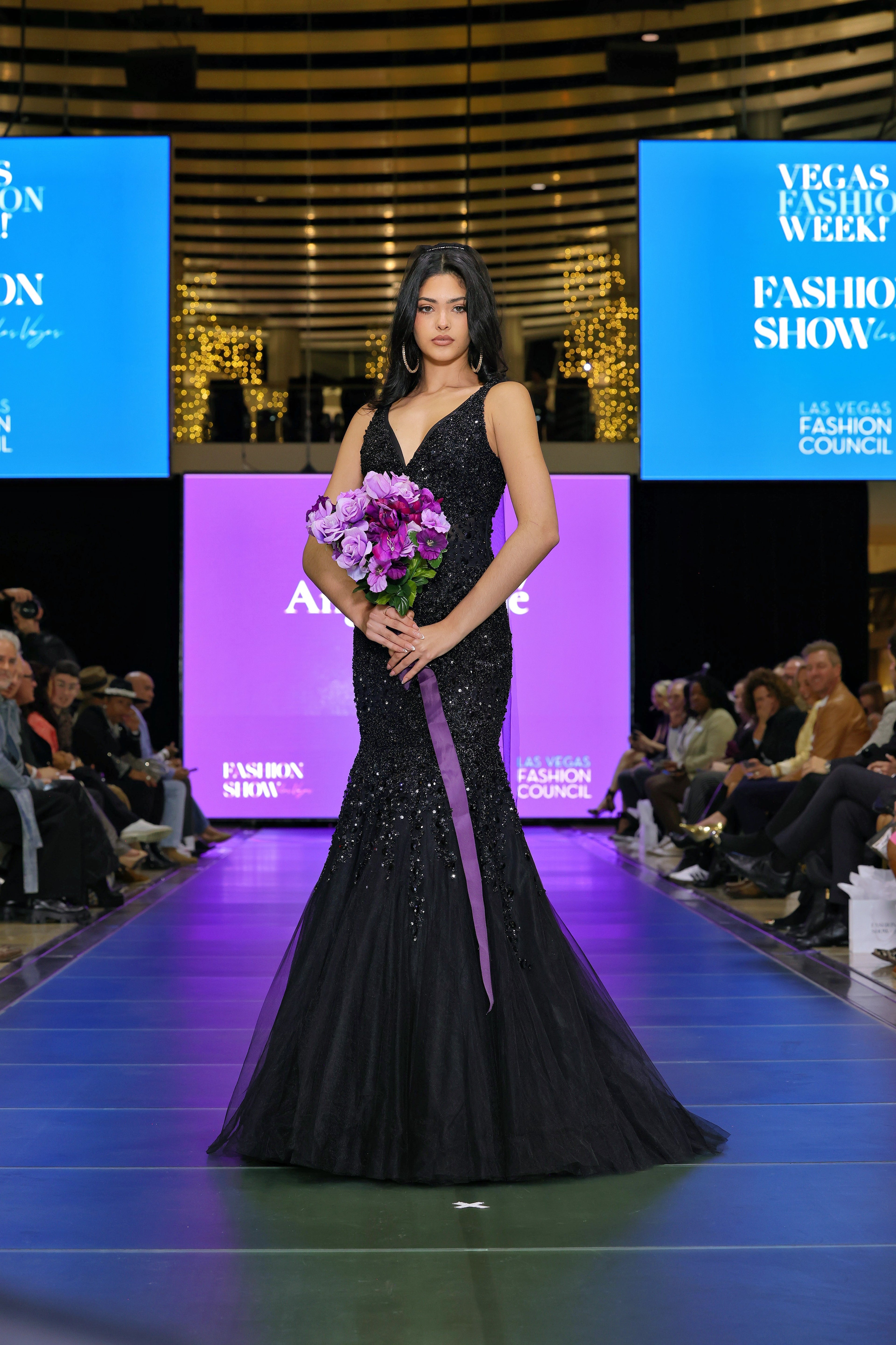 Woman in a black evening gown holding flowers on a runway with a blurred audience in the background.