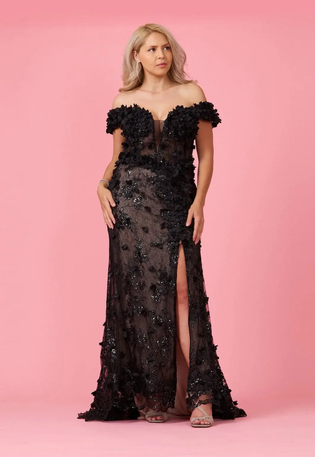 A model wearing an off-the-shoulder, full-length black gown with 3D floral corset detailing and a split on the left side.