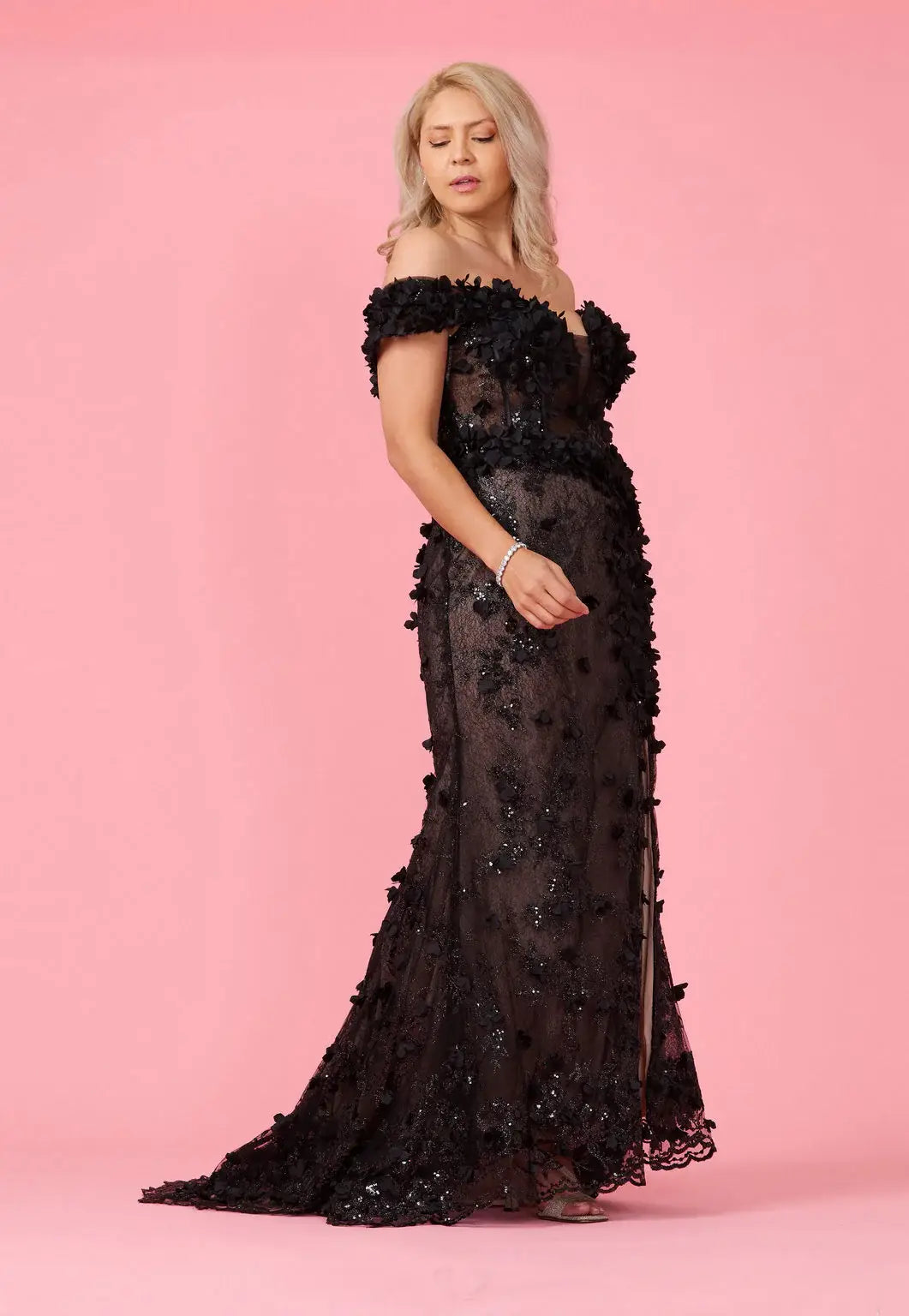 A model wearing an off-the-shoulder, full-length black gown with 3D floral corset detailing and a split on the left side.