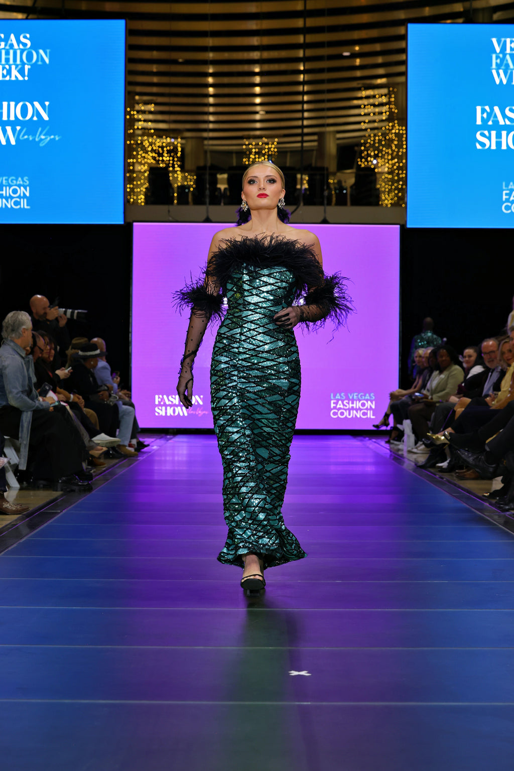 A model on a catwalk at a fashion show in a patterned gown with feathers.
