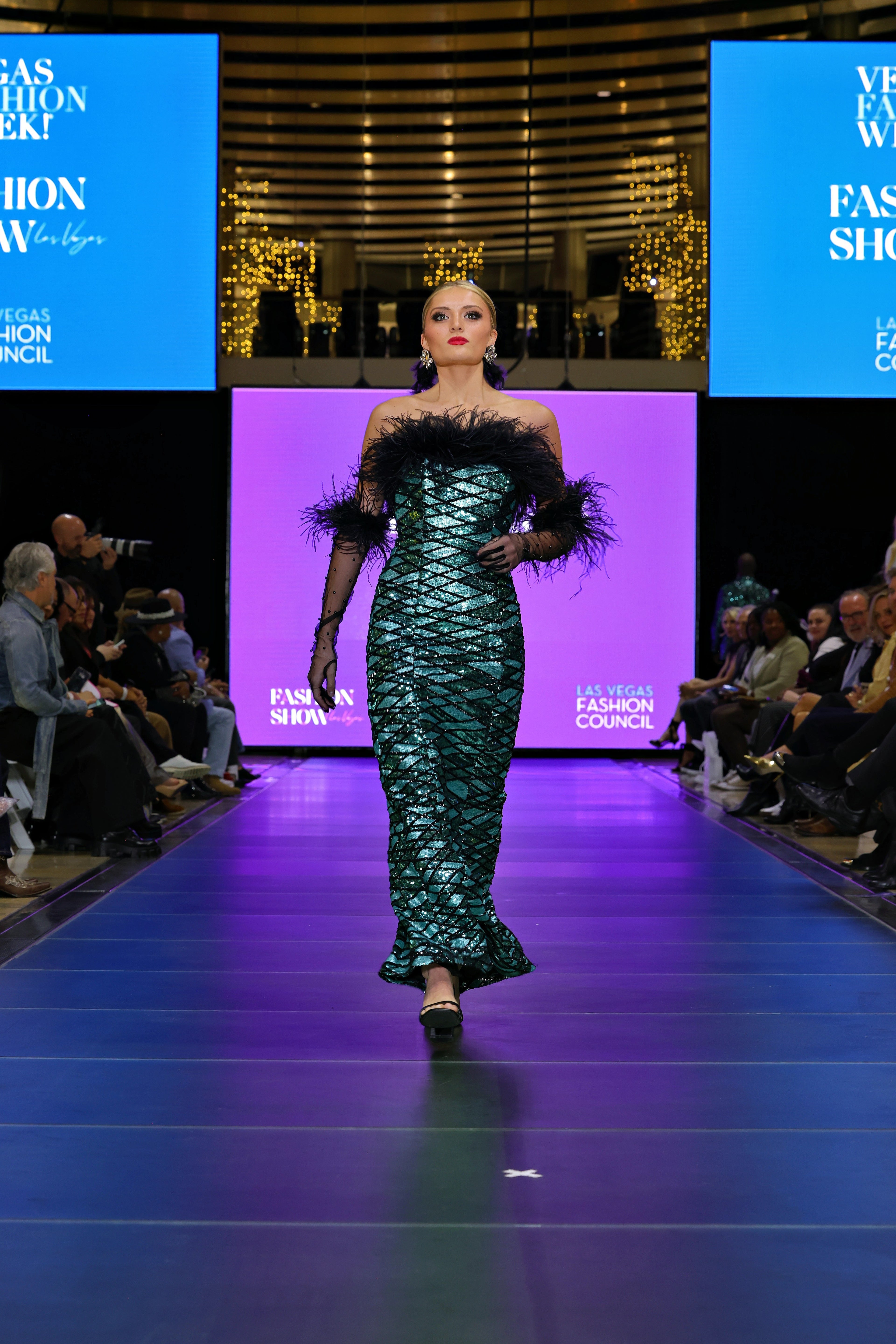 A model on a catwalk at a fashion show in a patterned gown with feathers.