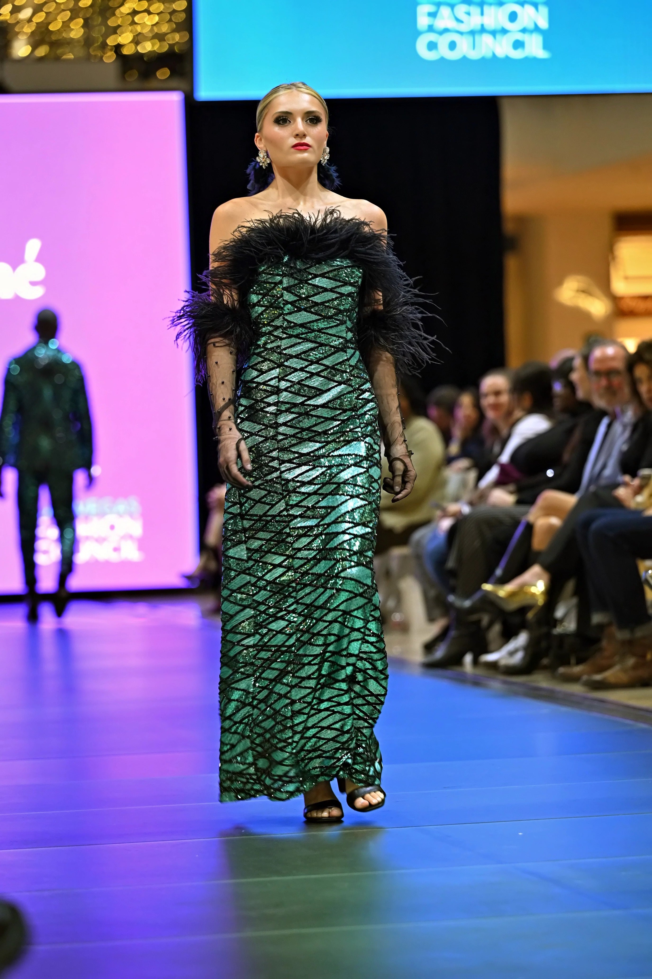 A model wearing a green sequin dress on a runway with a blurred audience in the background.